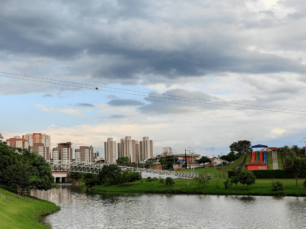 Lago de Indaiatuba cercado por gramados e árvores, com edifícios da cidade ao fundo sob um céu azul com nuvens.