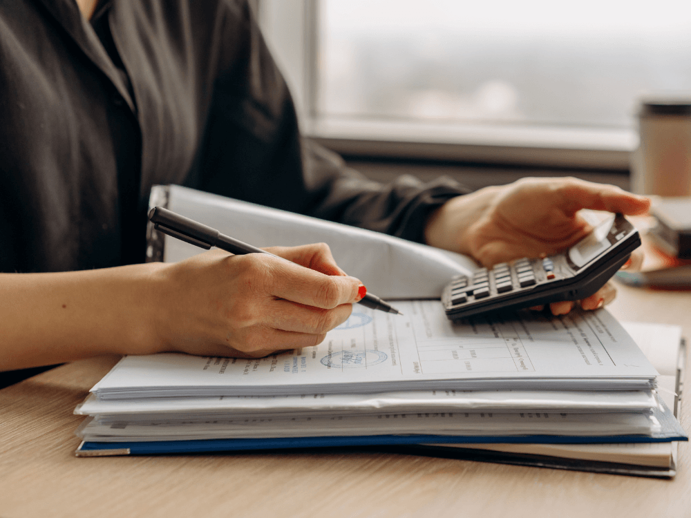 Pessoa utiliza uma calculadora e uma caneta sobre documentos organizados para calcular despesas relacionadas à compra de um imóvel.