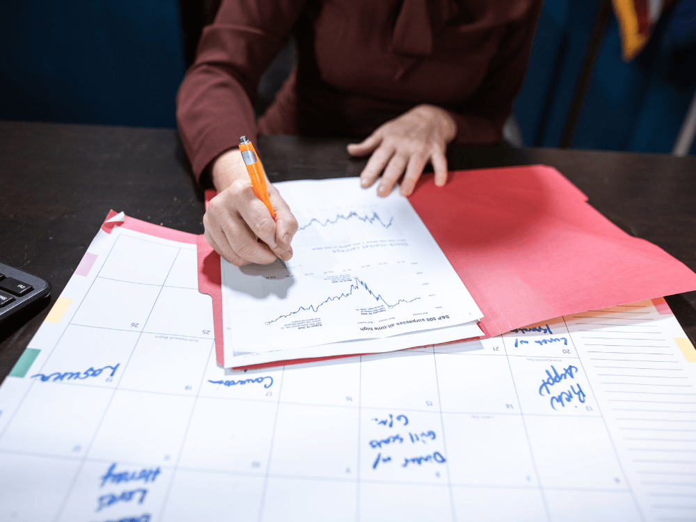 Pessoa preenche um documento com gráficos financeiros sobre uma mesa, ao lado de uma agenda e de uma pasta vermelha.
