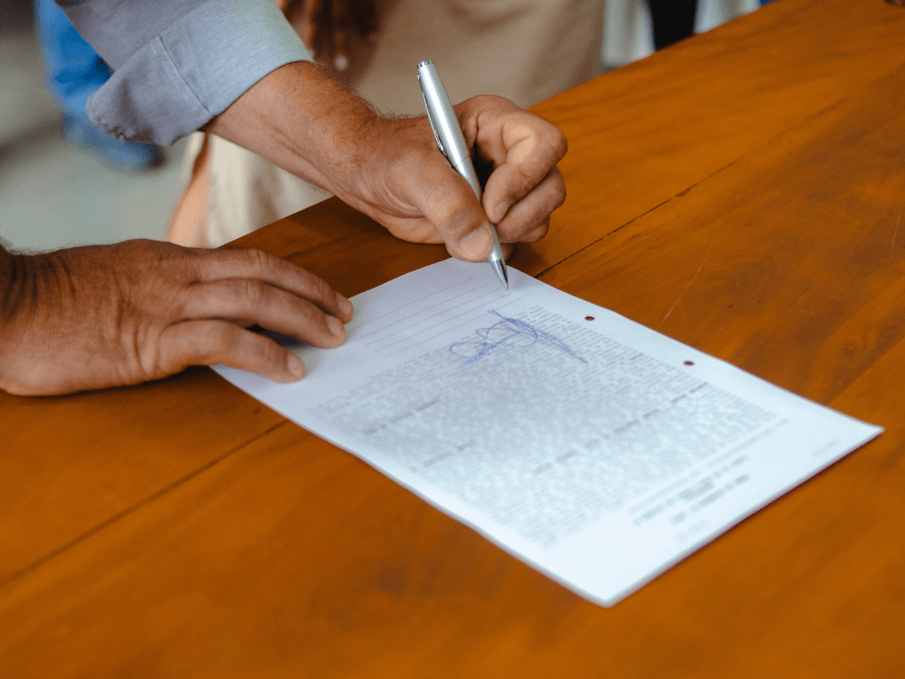 Pessoa assina um documento impresso sobre uma mesa de madeira, representando a formalização de um acordo de negociação imobiliária.
