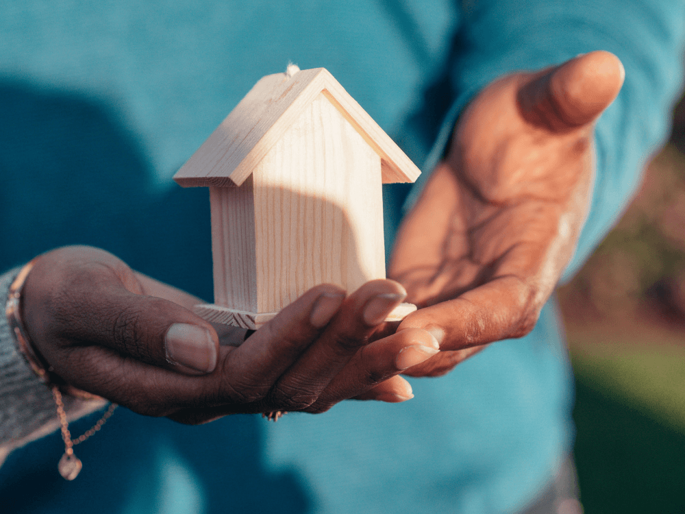 Mãos sustentam uma pequena casa de madeira, simbolizando a transferência de propriedade em uma negociação imobiliária.